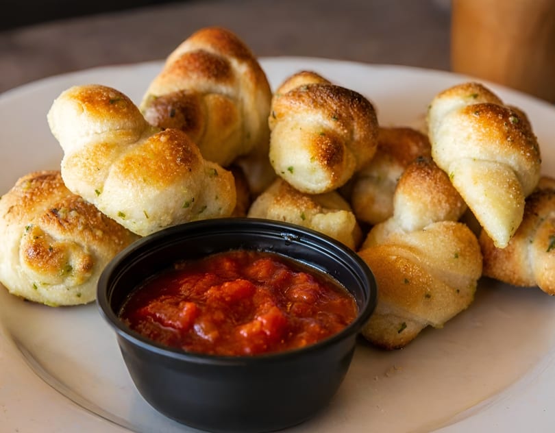 Golden brown garlic knots with herbs, served on a white plate with a side of marinara sauce.