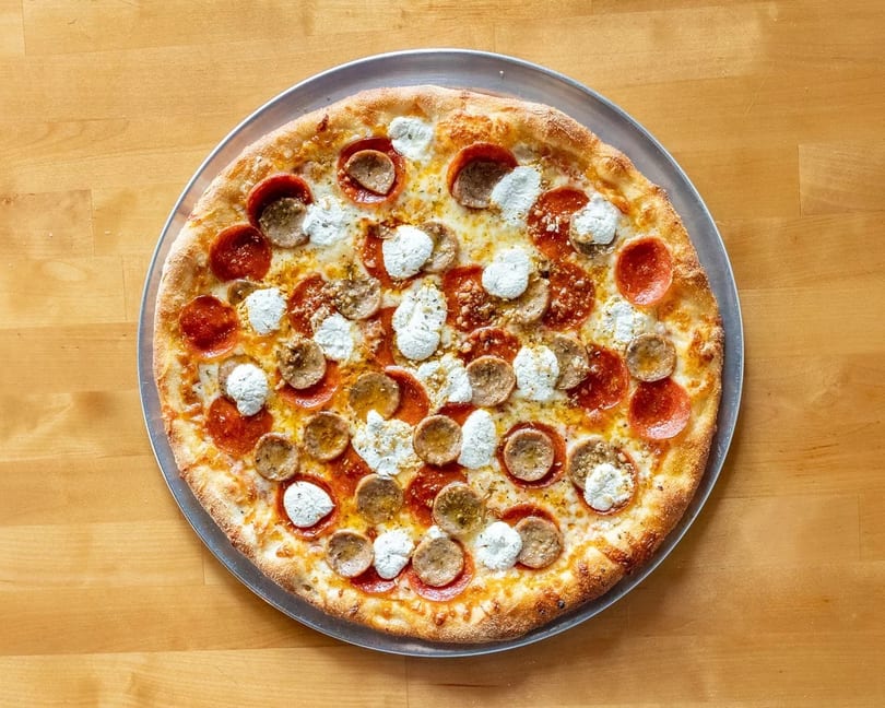 A whole pepperoni and sausage pizza with dollops of ricotta cheese on a metal tray.