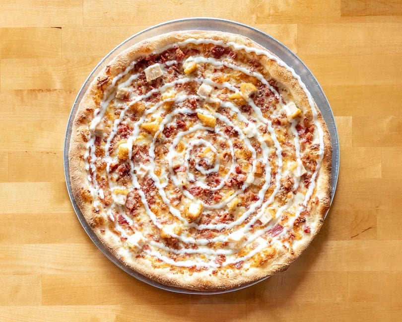 A whole pizza with chicken, bacon, and a spiral of white sauce on a wooden table.