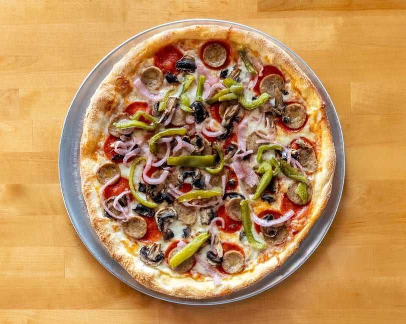 A whole pizza with pepperoni, sausage, green peppers, mushrooms, and red onions on a wooden table.